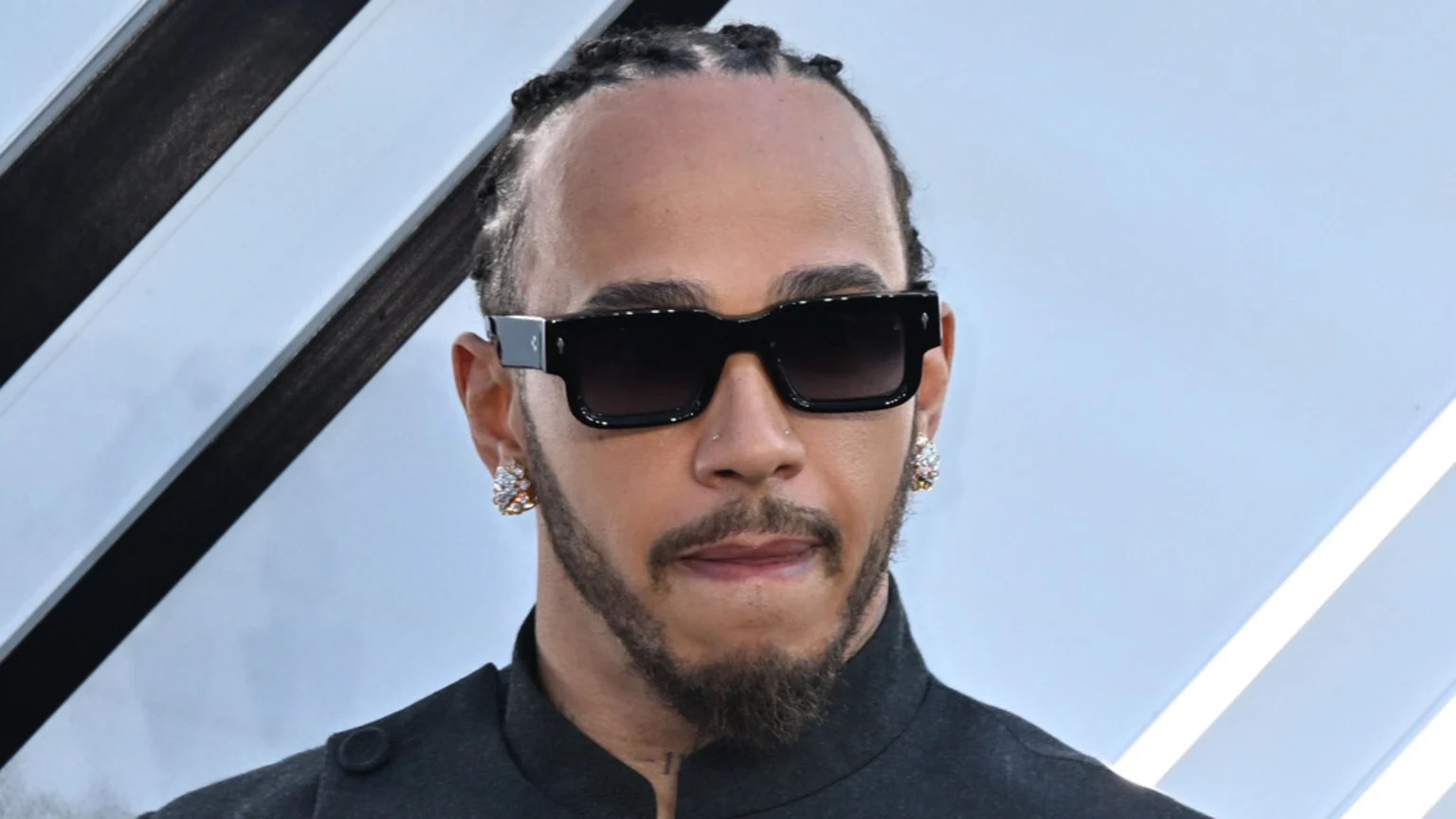 Close-up of Lewis Hamilton wearing black sunglasses, a beard, braided hair, and sparkling colorful earrings. He stands against a light blue and white background with diagonal black stripes.