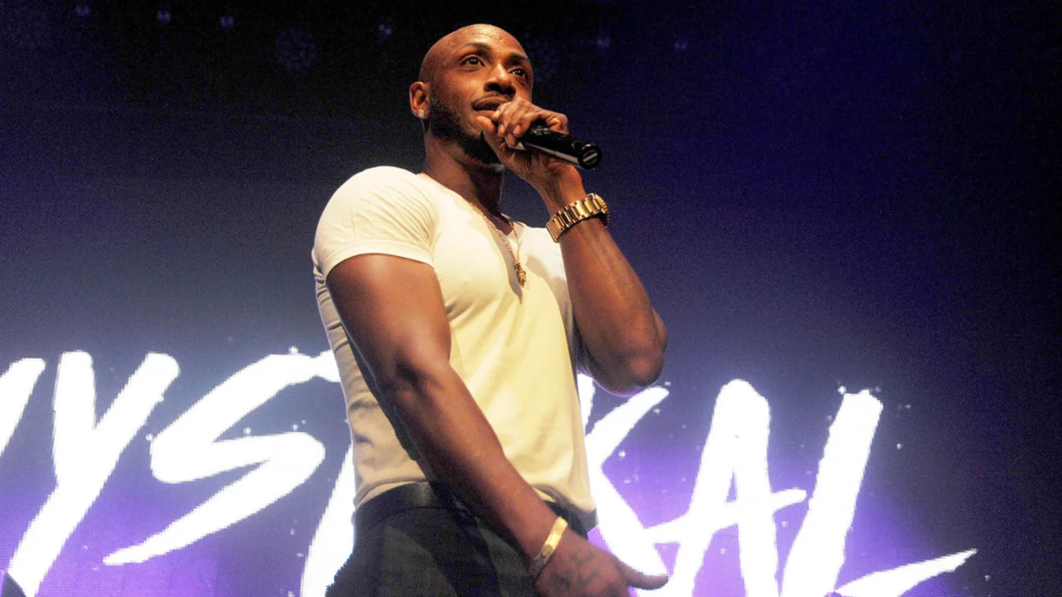 A bald, muscular man with a beard sings into a microphone on stage, wearing a white t-shirt and gold jewelry, with "MYSTIKAL" partially visible in white text in the background.