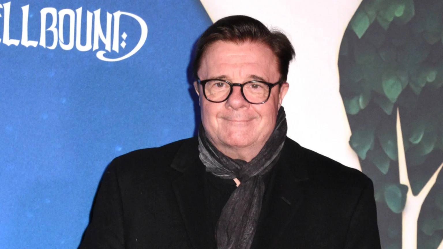 A smiling man with dark hair and glasses wears a black coat and a gray scarf against a split background of blue and white with green foliage.