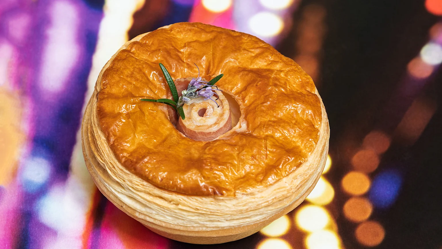 A golden-brown, flaky pastry pie with a rolled meat filling garnished with rosemary and purple flowers, set against a blurred, colorful bokeh background.