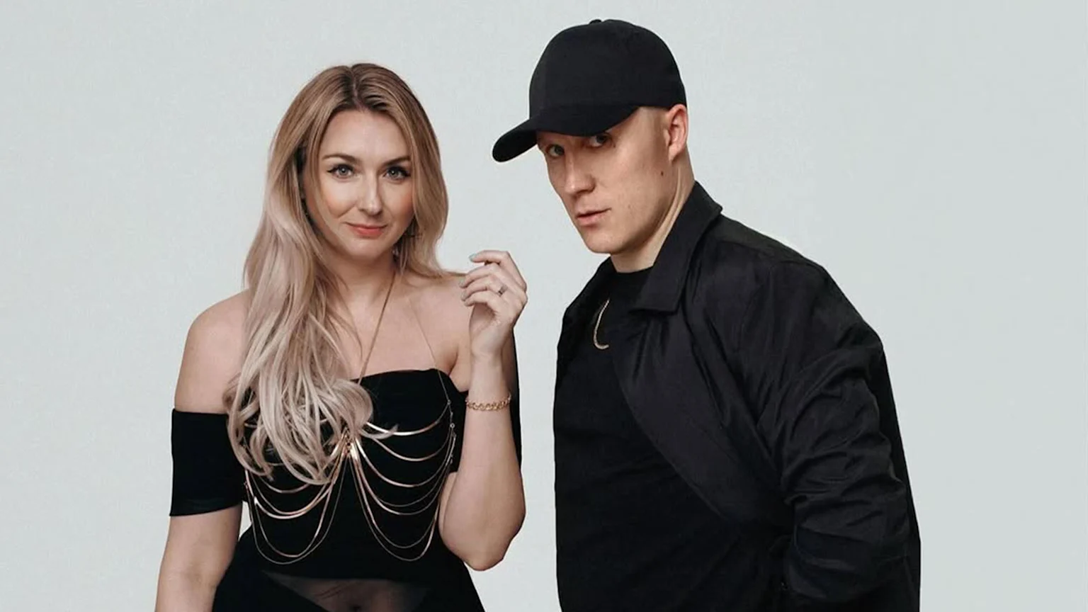 A blonde woman in a black off-shoulder top with a gold body chain and a man in a black cap and jacket with a gold necklace pose intently against a light background.
