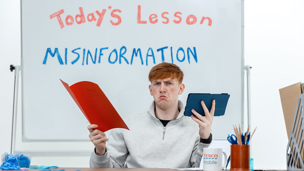 A young, red-haired man with a disgruntled expression holds a red folder and a blue tablet at a desk, with a whiteboard behind him displaying "Today's Lesson MISINFORMATION" and a Tesco Mobile mug.