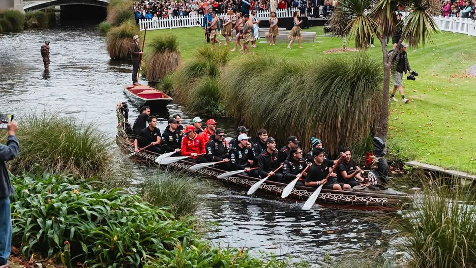 A group of men in a long, ornate wooden canoe paddles down a river past a standing figure in the water, as people in traditional attire and a crowd watch from the grassy bank.