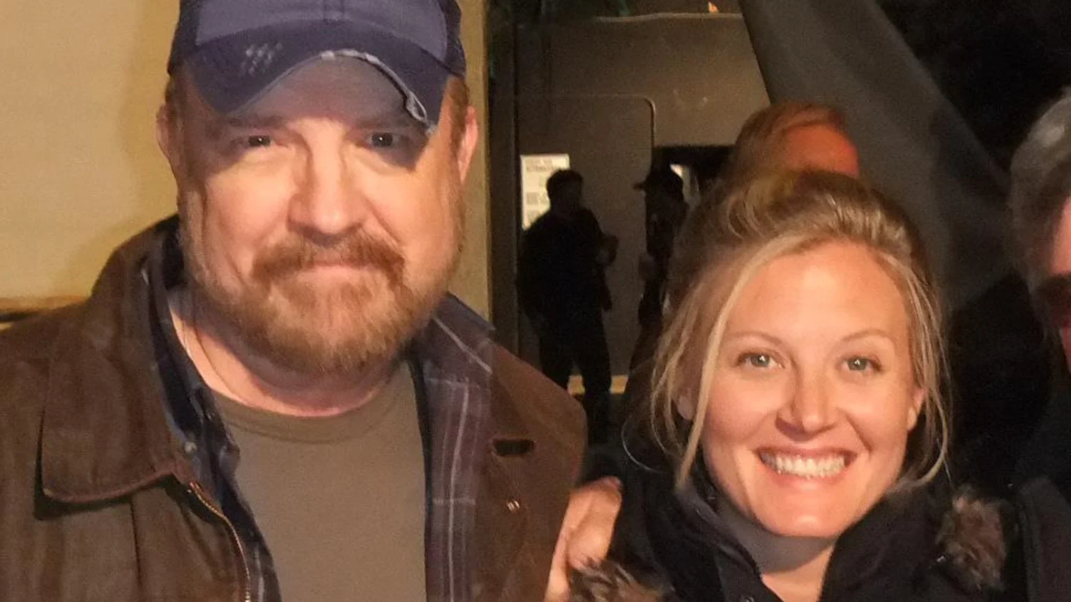 A smiling man with a beard and a baseball cap stands next to a smiling woman with blonde hair, both looking at the camera.