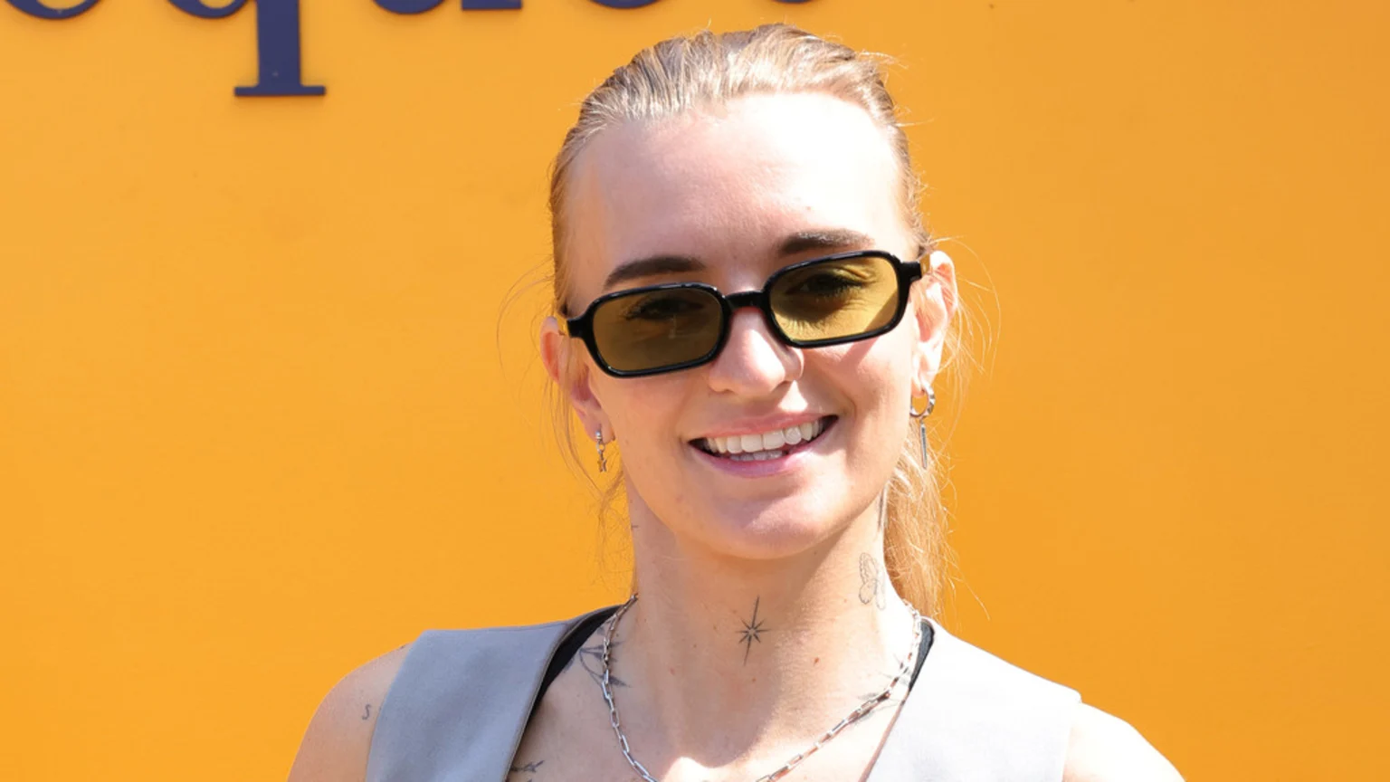 A smiling person with blonde hair, black sunglasses, and visible neck tattoos wears a silver necklace and a light top against an orange background with a partial blue letter at the top.