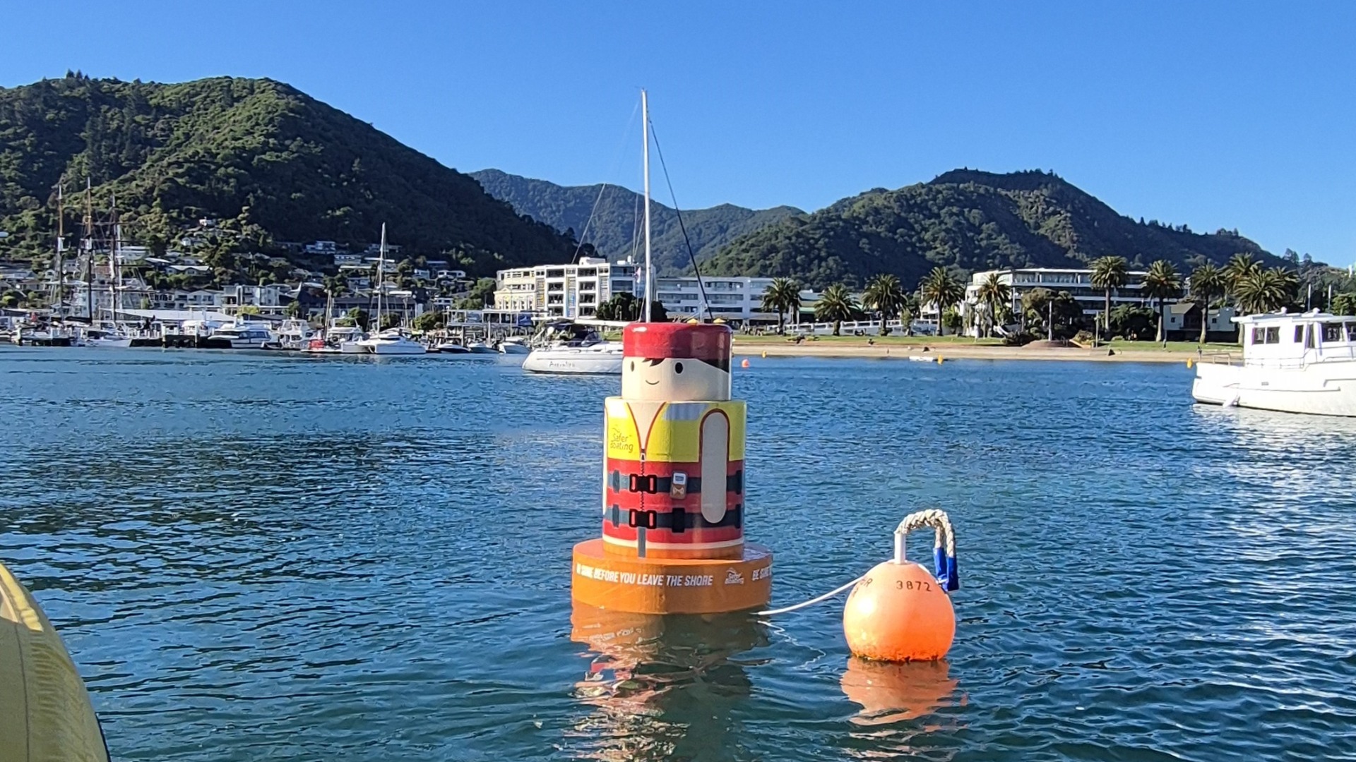 A cartoon-like buoy shaped like a person in a life jacket, marked "Safer Boating," floats in a bay with boats, buildings, palm trees, and green hills under a clear blue sky.