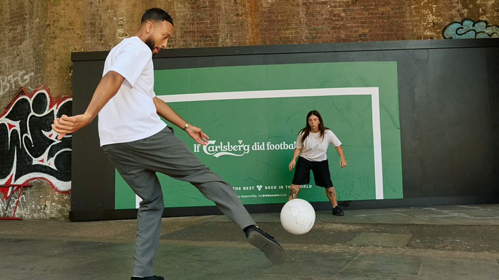 A man in a white t-shirt and grey pants kicks a white football towards a woman in a white t-shirt and black shorts, both playing in front of a green wall with "If Carlsberg did football" written on it.