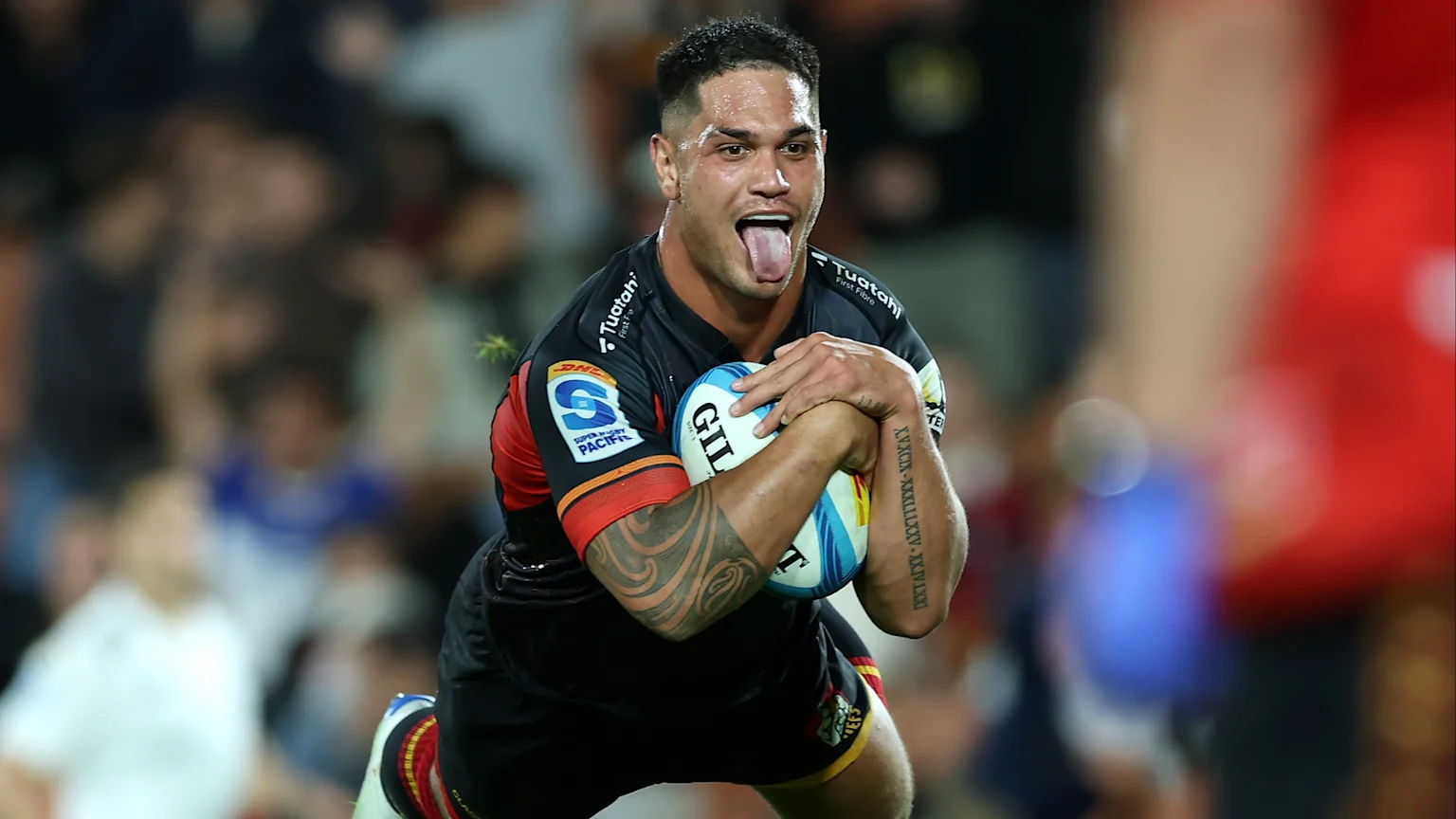 A male rugby player in a black and red Super Rugby Pacific jersey, with arm tattoos, holds a rugby ball and sticks out his tongue in front of a blurred crowd.
