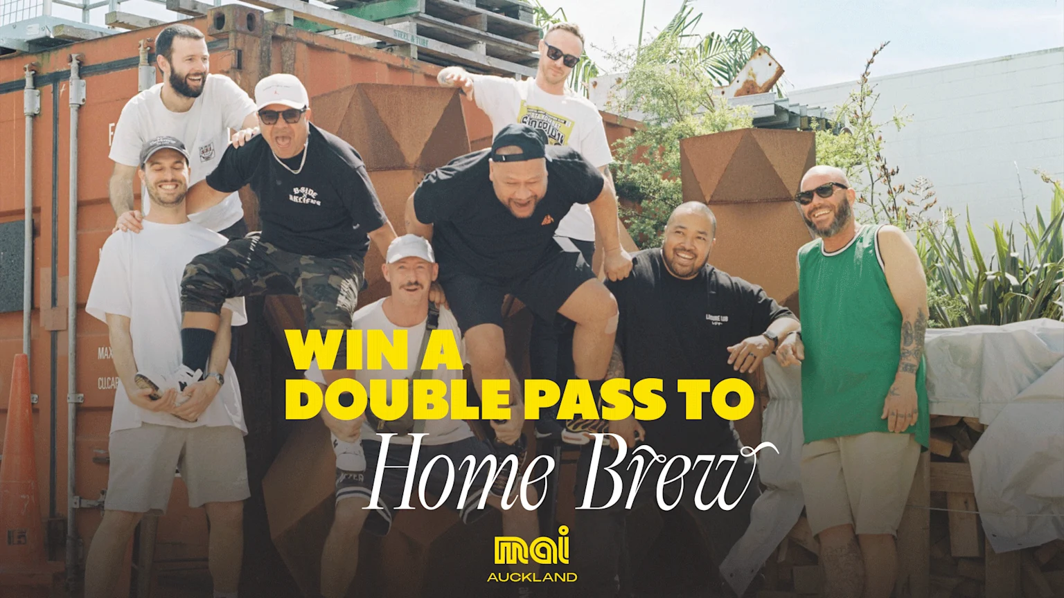 Eight diverse men smile playfully in a dynamic group pose outdoors, with shipping containers and plants in the background.