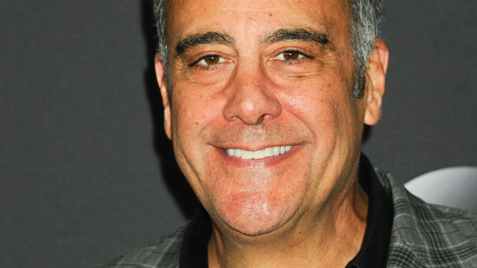 A close-up headshot of a smiling man with short, graying hair, brown eyes, and a patterned shirt, looking directly at the camera.