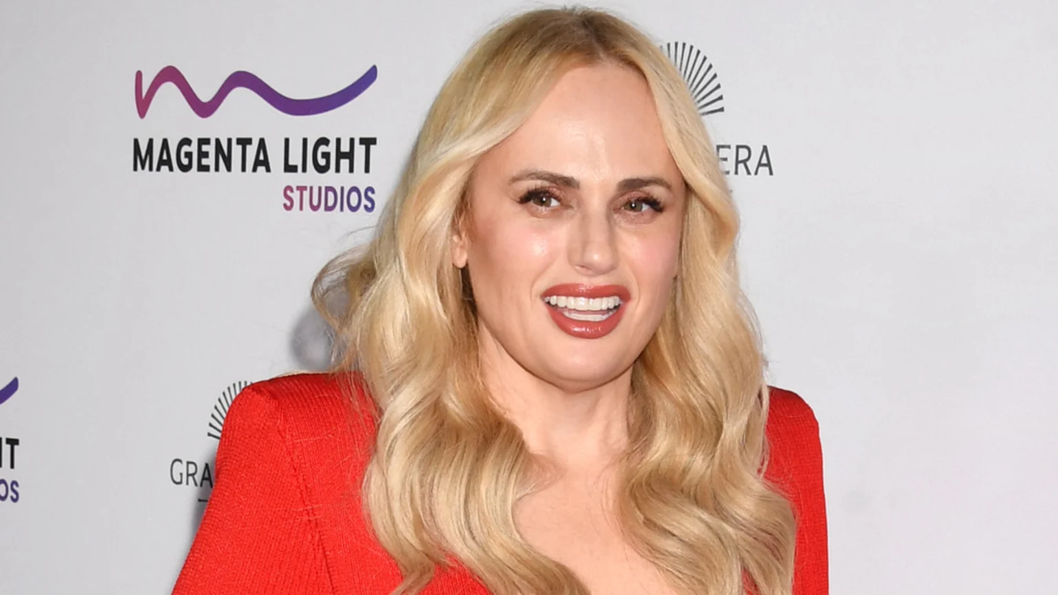Rebel Wilson, with long blonde hair and a red top, smiles at the camera, standing in front of a white backdrop with a "Magenta Light Studios" logo.