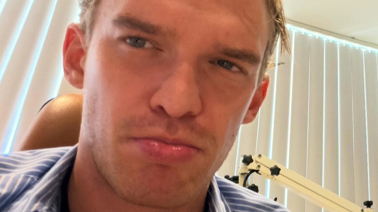 Close-up selfie of Cody Simpson with a serious expression, wearing a blue and white striped shirt, with vertical blinds visible in the background.