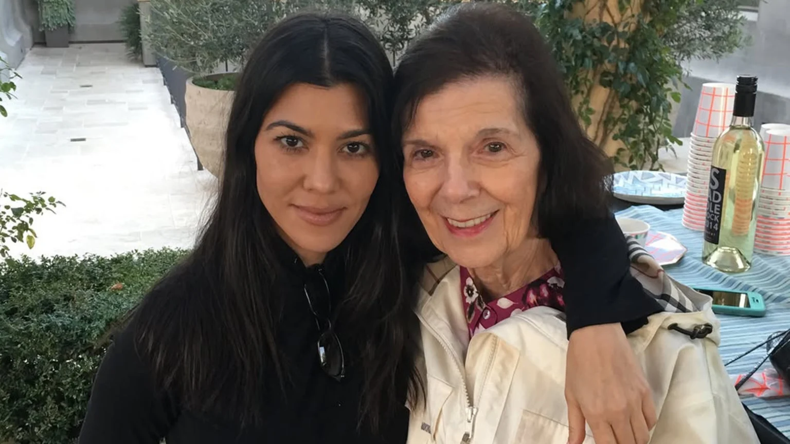 A smiling younger woman with dark hair embraces an older woman with dark, shorter hair in an outdoor setting with a table, wine, and cups in the background.