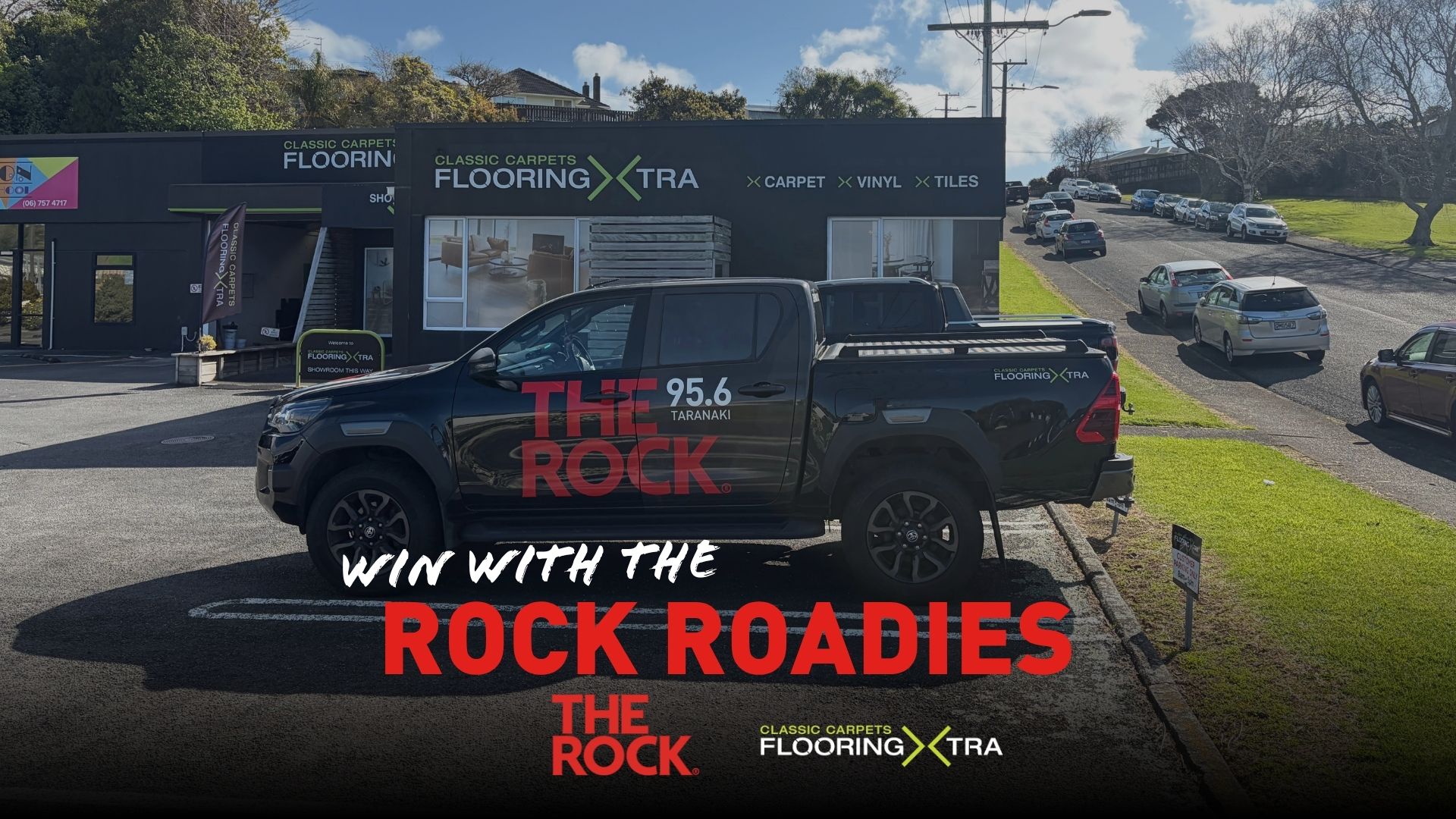 A black pickup truck branded "The Rock 95.6 Taranaki" is parked outside "Classic Carpets Flooring Xtra" store, with "Win with The Rock Roadies" text overlay.