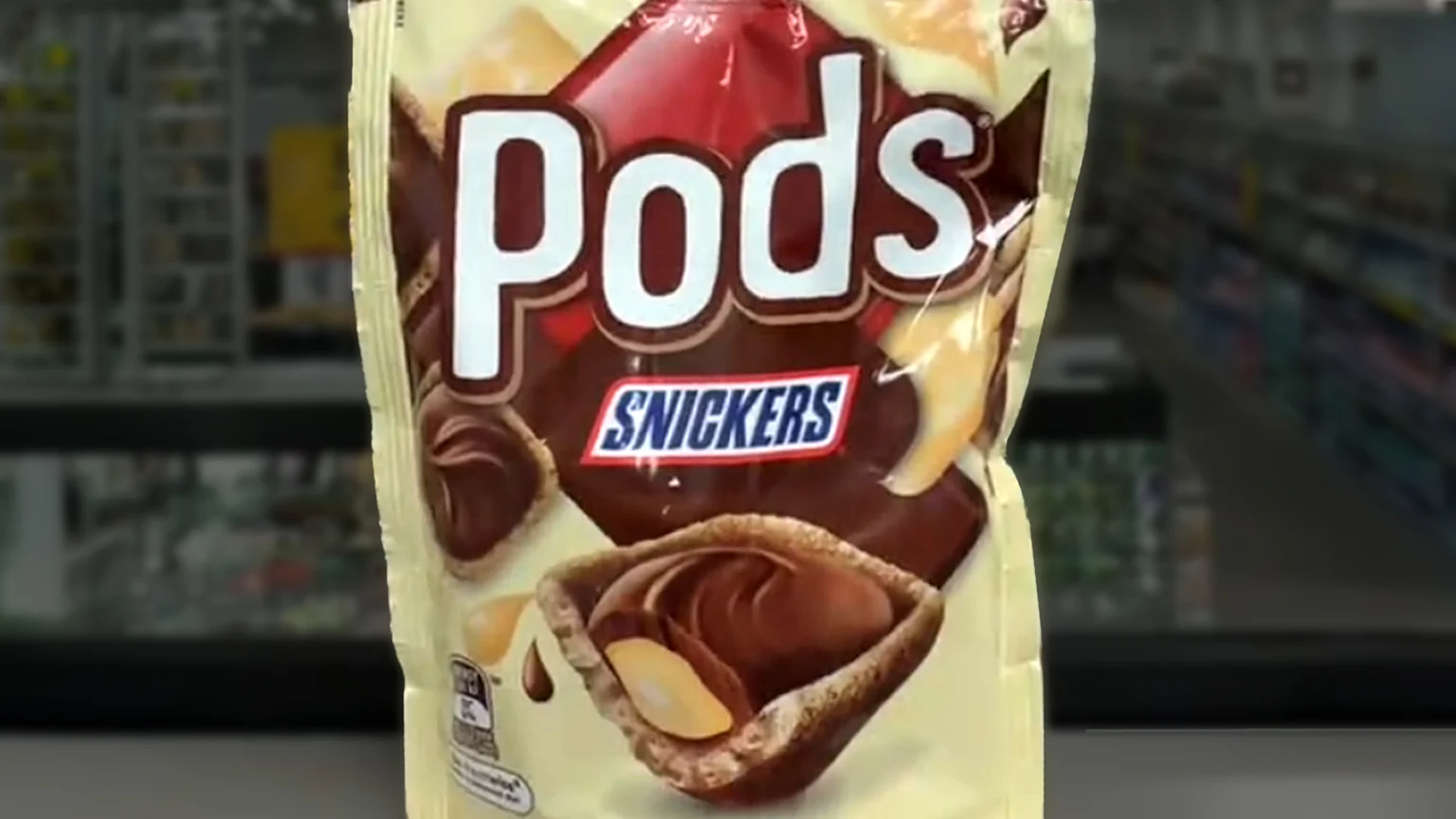A light yellow and brown bag of Pods Snickers snacks features biscuit cups filled with chocolate, caramel, and peanuts, displayed against a blurred supermarket aisle.
