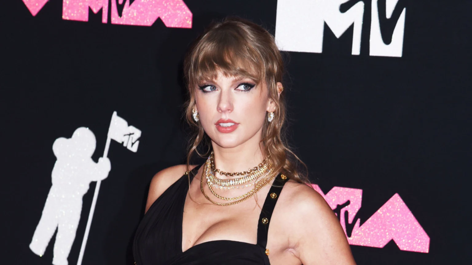 Taylor Swift in a black dress and layered gold necklaces, posing against a black background featuring white and pink glitter MTV logos and the white MTV astronaut figure.