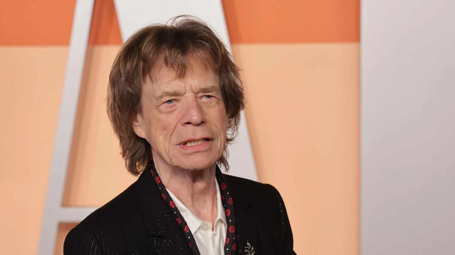 A close-up of Mick Jagger, an older man with brown hair, a furrowed brow, and his mouth slightly open, wearing a dark jacket and a red-and-black patterned scarf, against a blurred background.