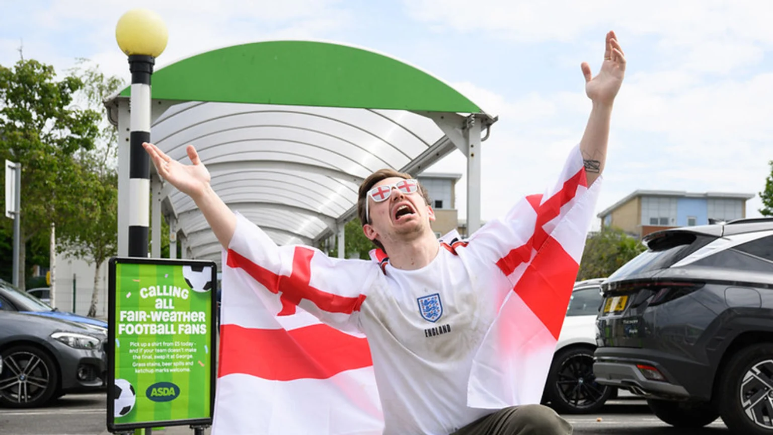 A man in an England football shirt and flag, wearing cross-shaped sunglasses, cheers with arms raised next to an Asda sign advertising a "fair-weather football fans" promotion.