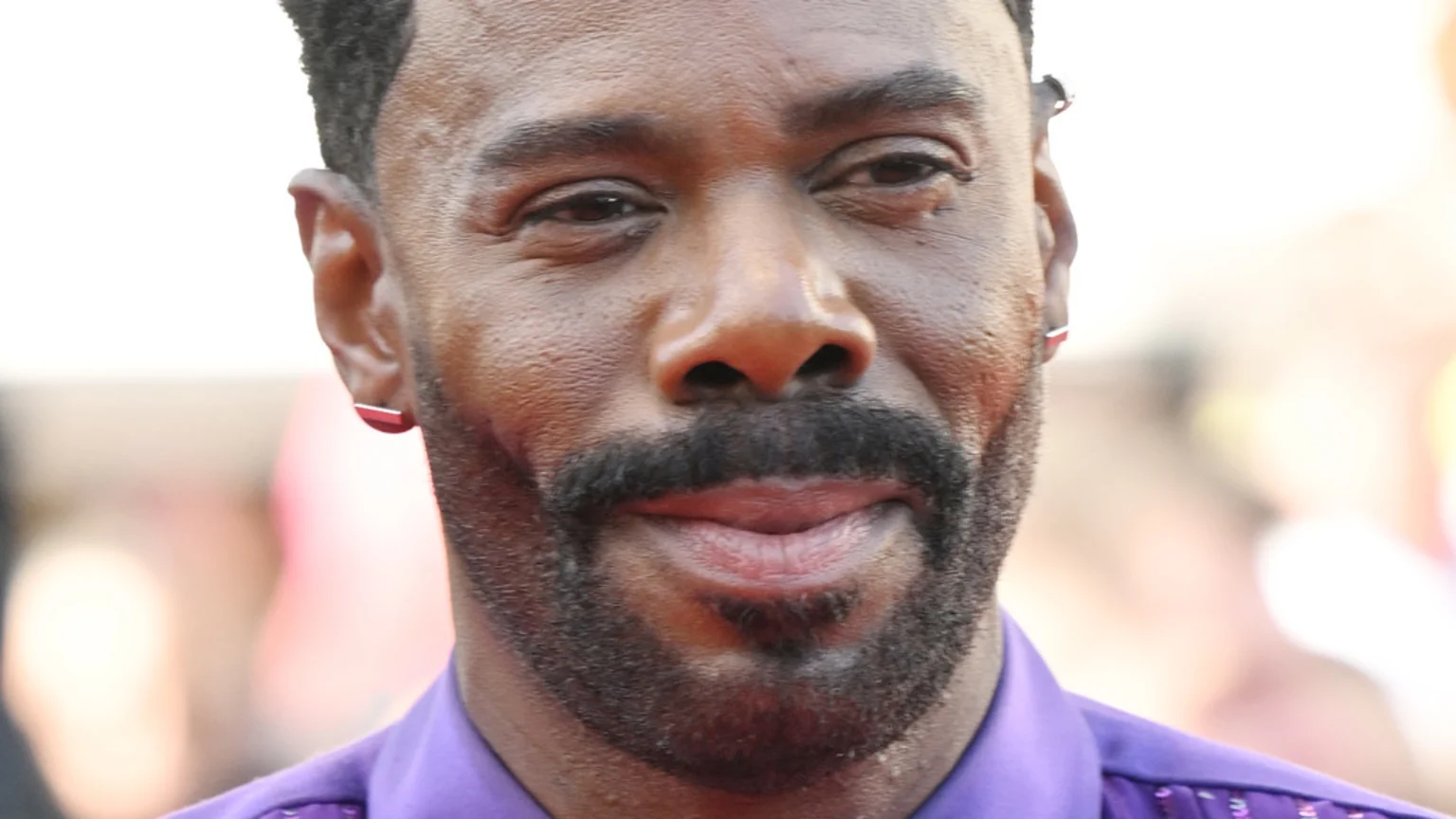 Close-up of actor Colman Domingo smiling, with a beard and mustache, wearing a purple shirt and reddish-pink stud earrings.