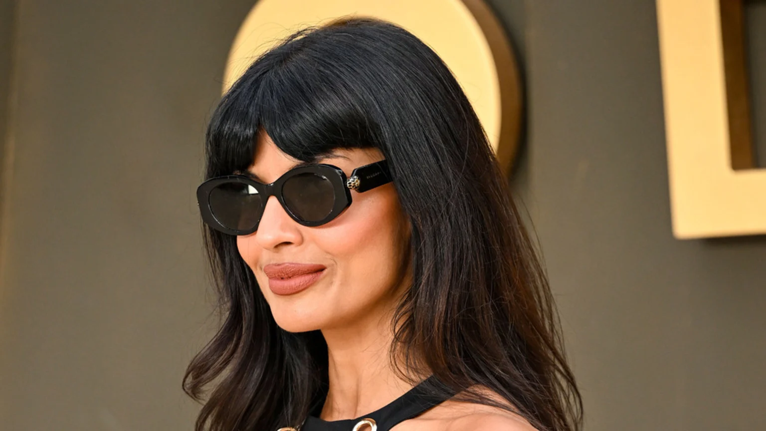 Jameela Jamil, with dark hair and bangs, smiles subtly while wearing black sunglasses and a dark top.