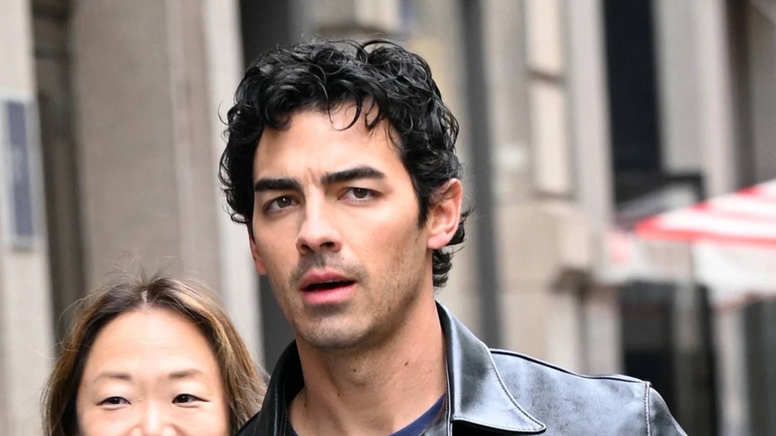 Joe Jonas, with dark curly hair and a leather jacket, looks forward with a slightly open mouth; a person with light brown hair and Asian features is partially visible behind him.