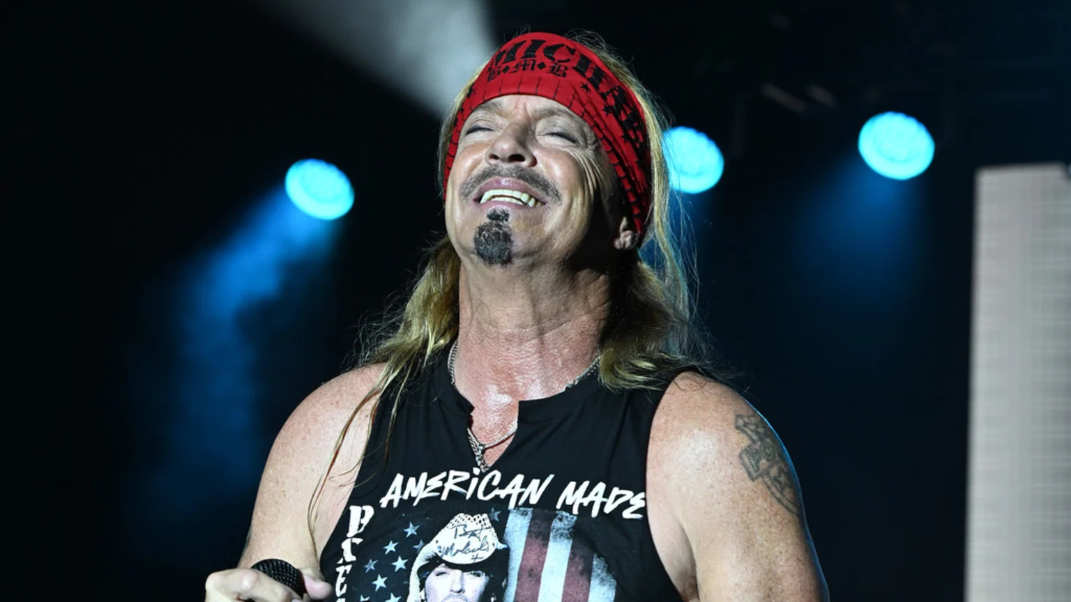 A man with blonde hair and a goatee, wearing a red bandana and an "AMERICAN MADE" tank top, sings into a microphone on a stage with blue lights.