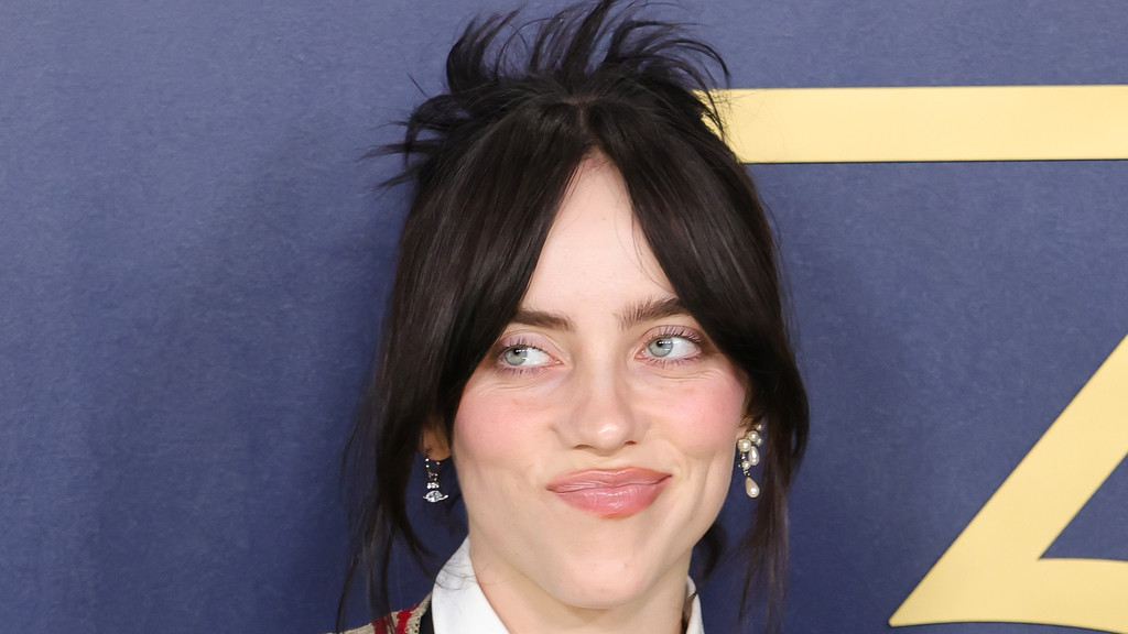 Close-up portrait of Billie Eilish with dark hair styled in bangs and a messy bun, looking left with a slight smirk, wearing pearl earrings against a dark blue background with a gold stripe.