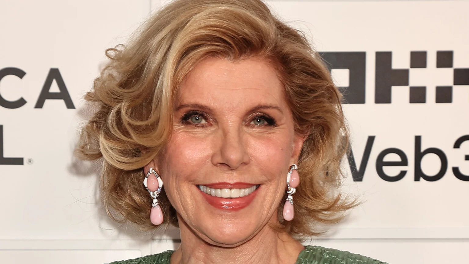 A close-up shot of actress Christine Baranski smiling, with blonde hair, light eyes, and pink drop earrings, against a white background with partial black logos.