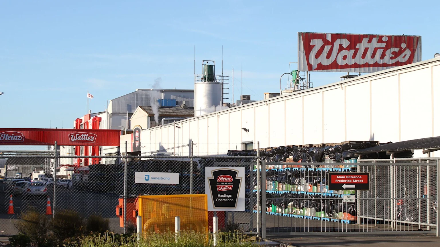 A large Wattie's factory building is seen behind a chain-link fence, with a parking lot full of cars in front under a clear blue sky.