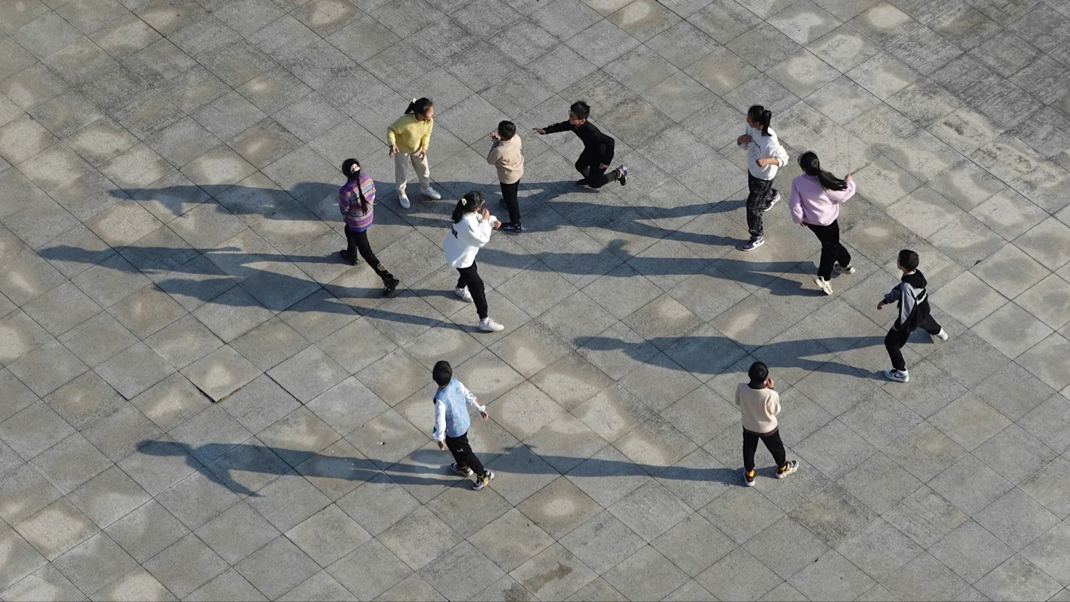 An overhead view shows a group of children actively playing on a grey tiled outdoor surface, with their long shadows cast behind them.