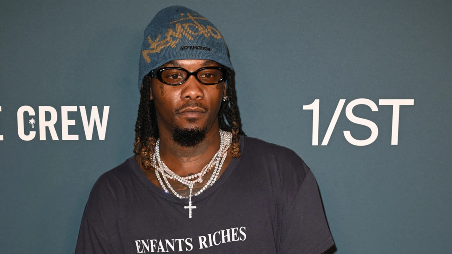 Offset, a man with dreadlocks and a beard, looks directly at the camera, wearing a blue beanie, dark glasses, a dark t-shirt, and layered diamond necklaces, including a cross pendant, against a teal background with white text.