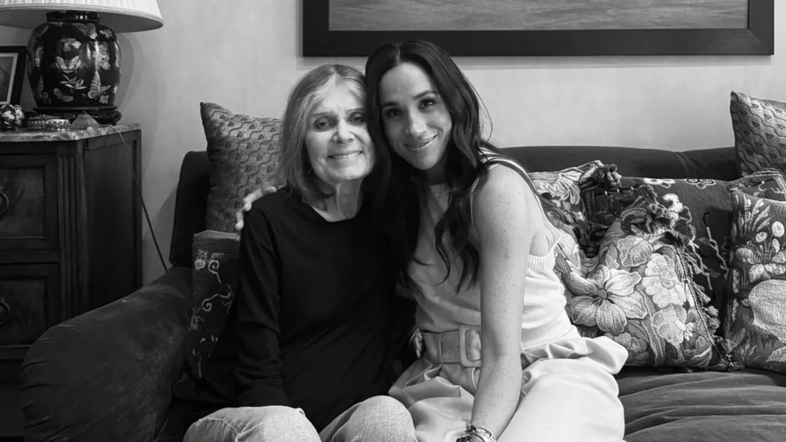 In a black and white image, Meghan Markle and Gloria Steinem sit closely on a couch, both smiling warmly at the camera, with Steinem's arm gently behind Meghan.