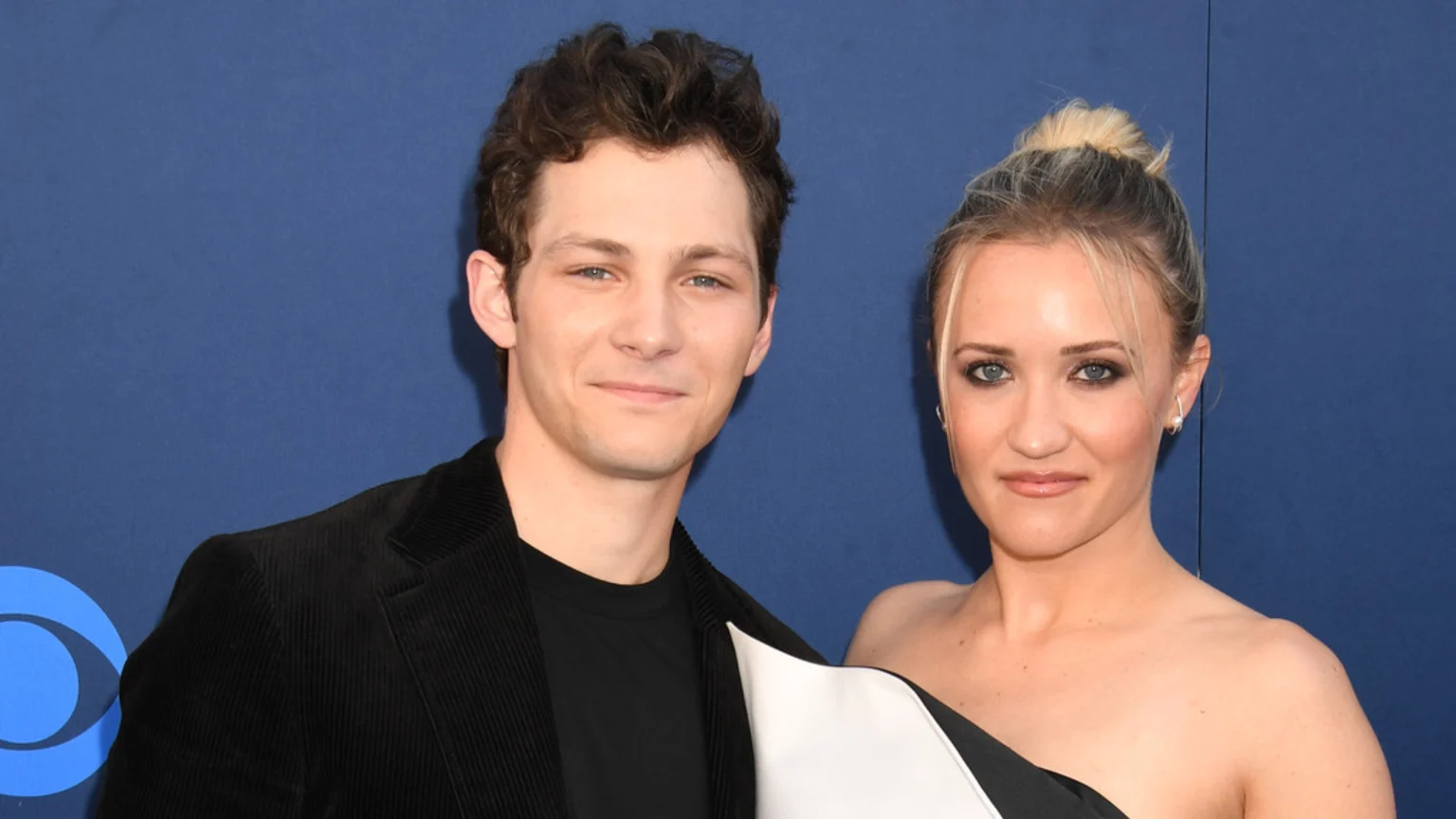 A smiling man with curly brown hair in a black jacket stands next to a smiling woman with blonde hair pulled up, wearing a black and white one-shoulder dress, against a blue background with a partial CBS logo.