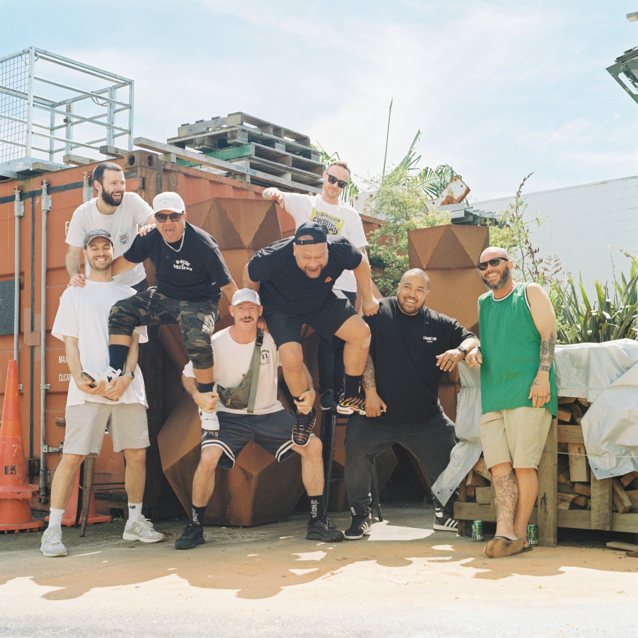 Eight smiling men of varying ethnicities pose in a playful human pyramid outdoors, dressed casually against a backdrop of shipping containers, pallets, and greenery under a bright sky.