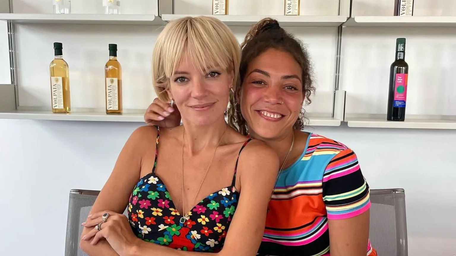 Lily Allen, a blonde woman in a floral top, smiles next to a curly-haired woman in a striped top, with bottles on white shelves in the background.