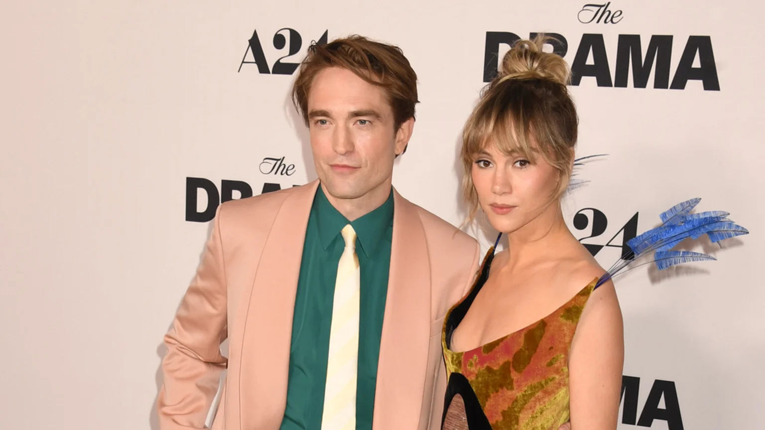 Robert Pattinson in a peach suit and green shirt stands next to Suki Waterhouse in a patterned dress with blue shoulder details, against A24 and The DRAMA logos.