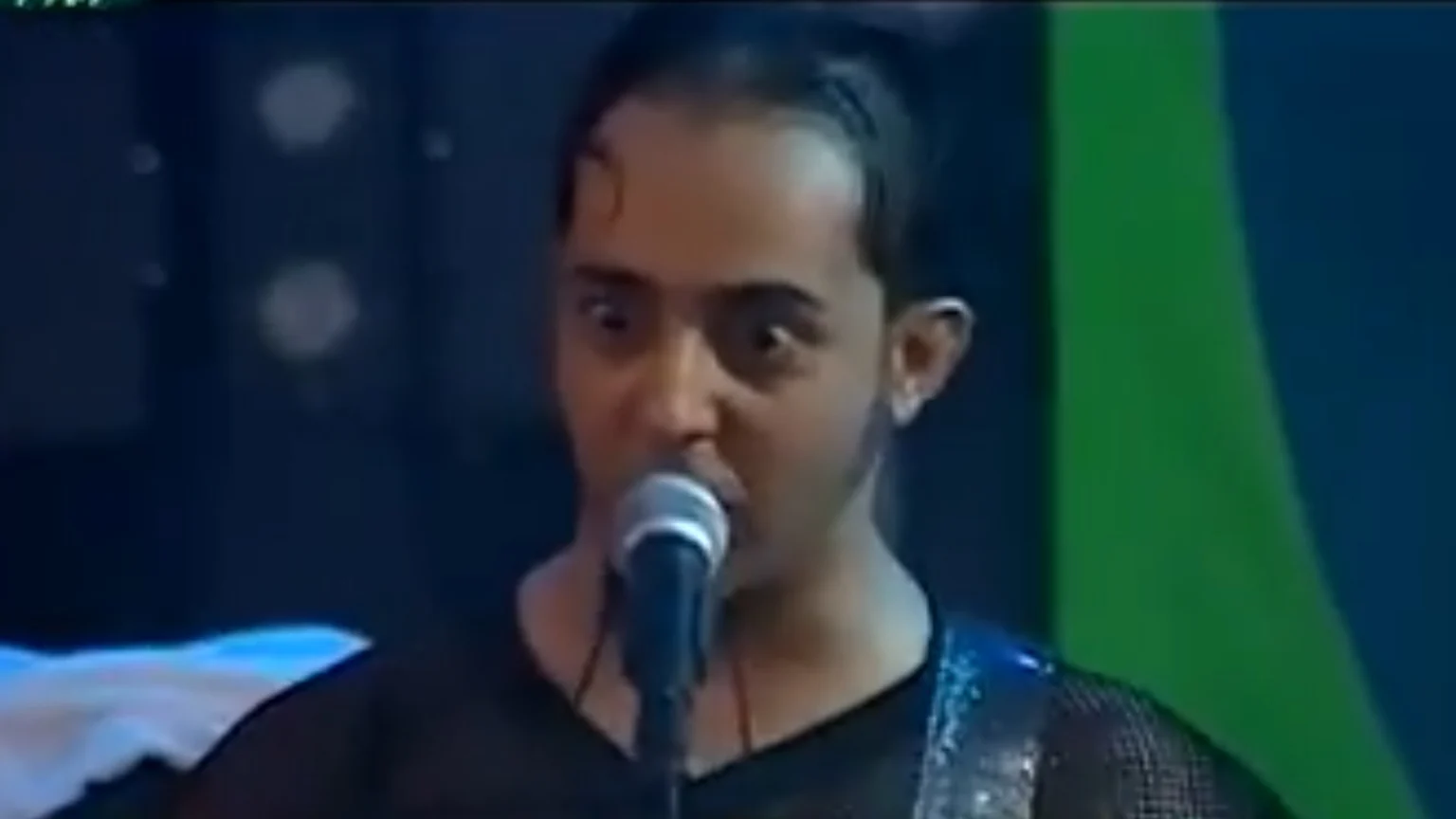 A man with wide eyes and a receding hairline sings intensely into a microphone on stage, wearing a dark shirt with a guitar strap, against a dark blue and green background with blurry lights.