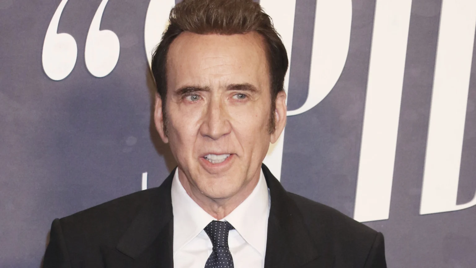 Nicolas Cage, wearing a black suit and a polka dot tie, looks directly at the camera with his mouth slightly open, against a blurred background with large white graphic elements.