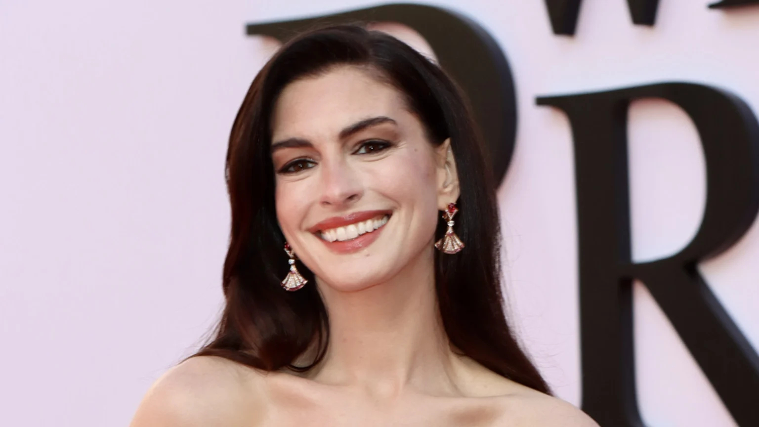 Close-up of Anne Hathaway smiling, with dark hair and red and gold earrings, against a light background with dark letters.