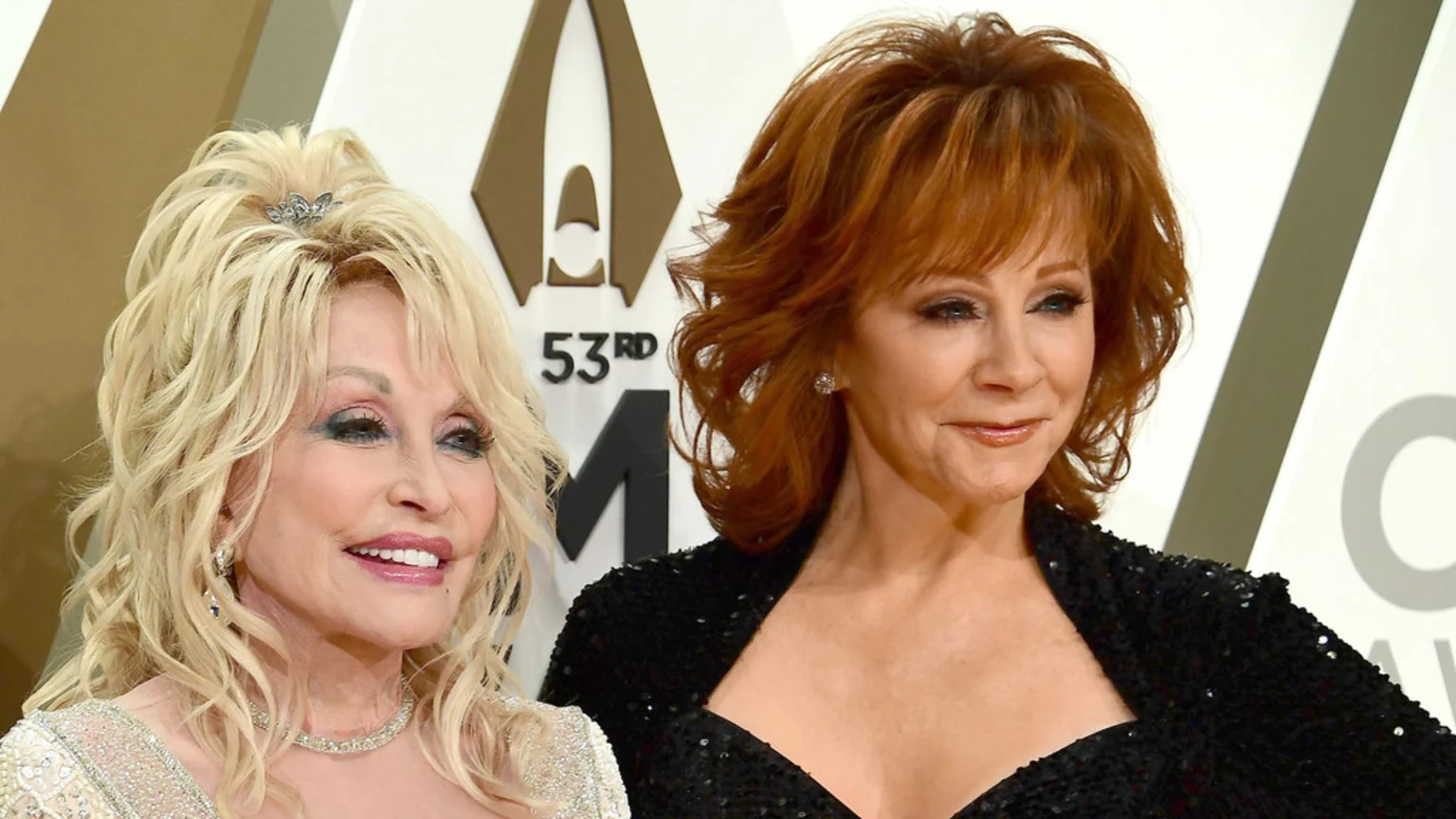 Two smiling country music legends, Dolly Parton with blonde hair and a white beaded dress on the left, and Reba McEntire with red hair and a black sequined top on the right, pose together.