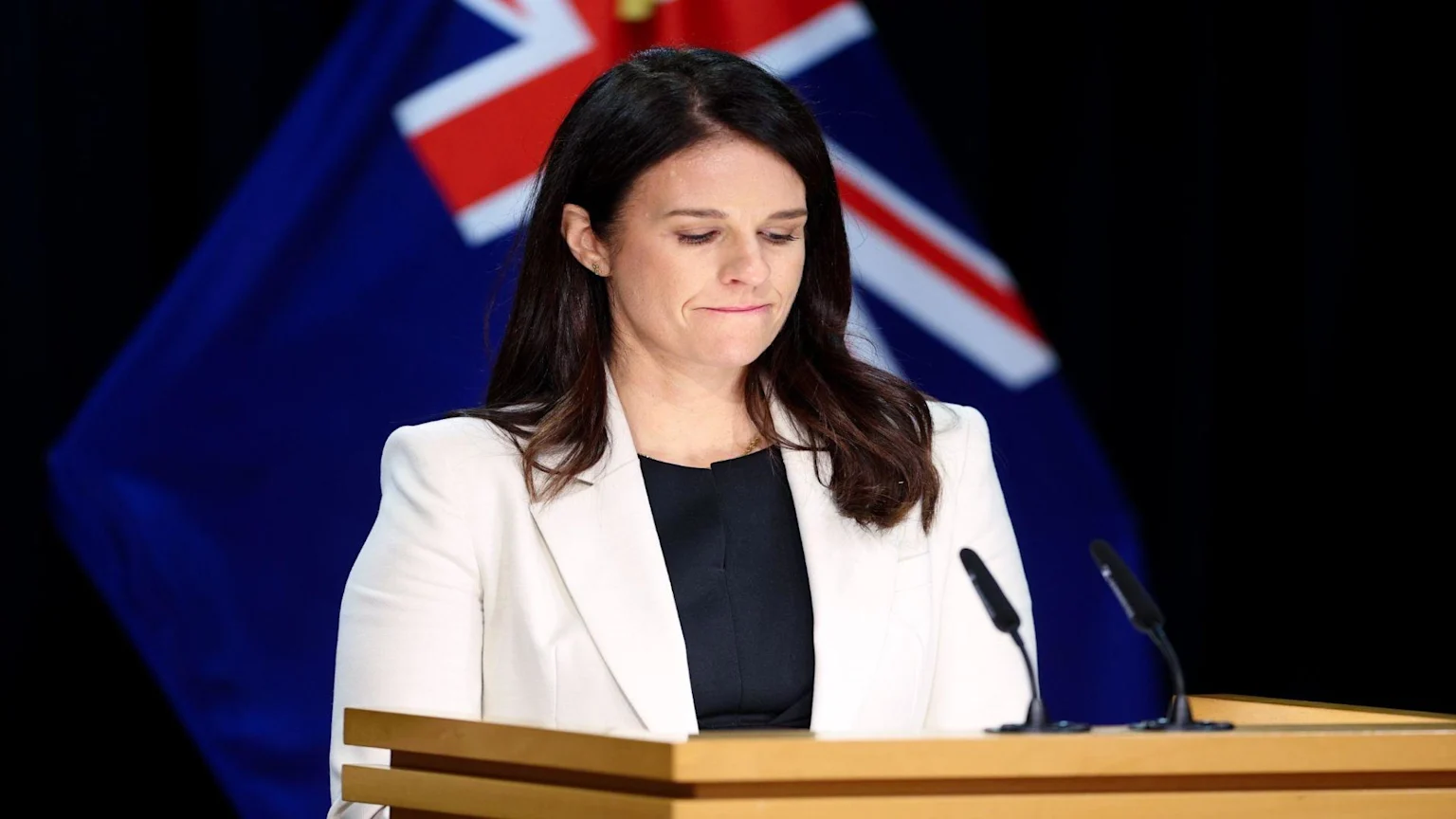 A woman with dark hair in a white blazer and black top stands behind a podium with microphones, looking down with a slight frown, against a backdrop of a blue flag.