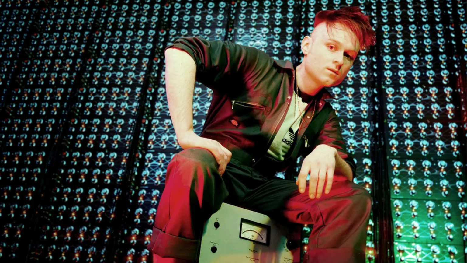 A young man with styled red hair wears a dark zippered jumpsuit and a light t-shirt featuring a black Adidas logo. He sits on a light grey vintage electronic device with analog meter and control knobs. The background displays an illuminated circuit board pattern with blue-green and red-orange circular elements. Dramatic red and blue-green lighting highlights the scene in a studio environment.