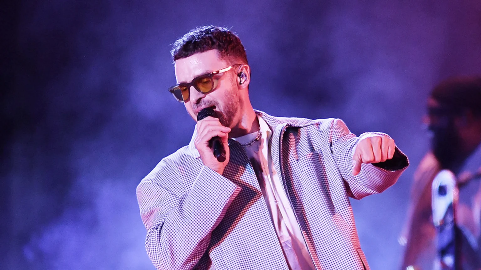 Justin Timberlake performs on stage, wearing yellow-tinted sunglasses, a checkered jacket, and holding a microphone, with his right arm extended.