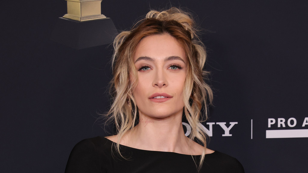 Paris Jackson with blonde hair pulled up, wearing a black top, looks at the camera against a dark background with faint logos.