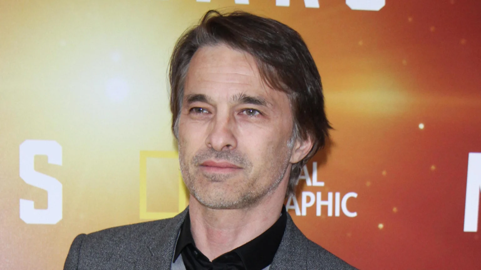 Close-up of actor Olivier Martinez, a man with dark hair and stubble, wearing a dark suit jacket over a dark shirt, against a blurry yellow and orange background with "NATIONAL GEOGRAPHIC" text.