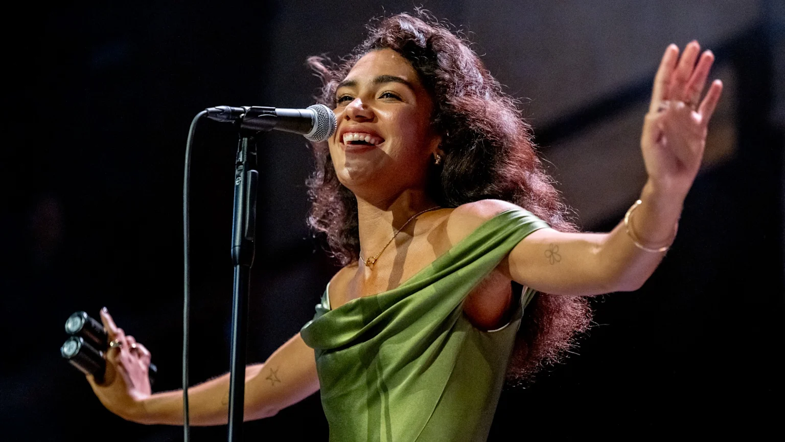 Olivia Dean, with curly hair, smiles widely while singing into a microphone on stage, wearing a green off-the-shoulder dress, with her right hand raised and holding two small instruments in her left.
