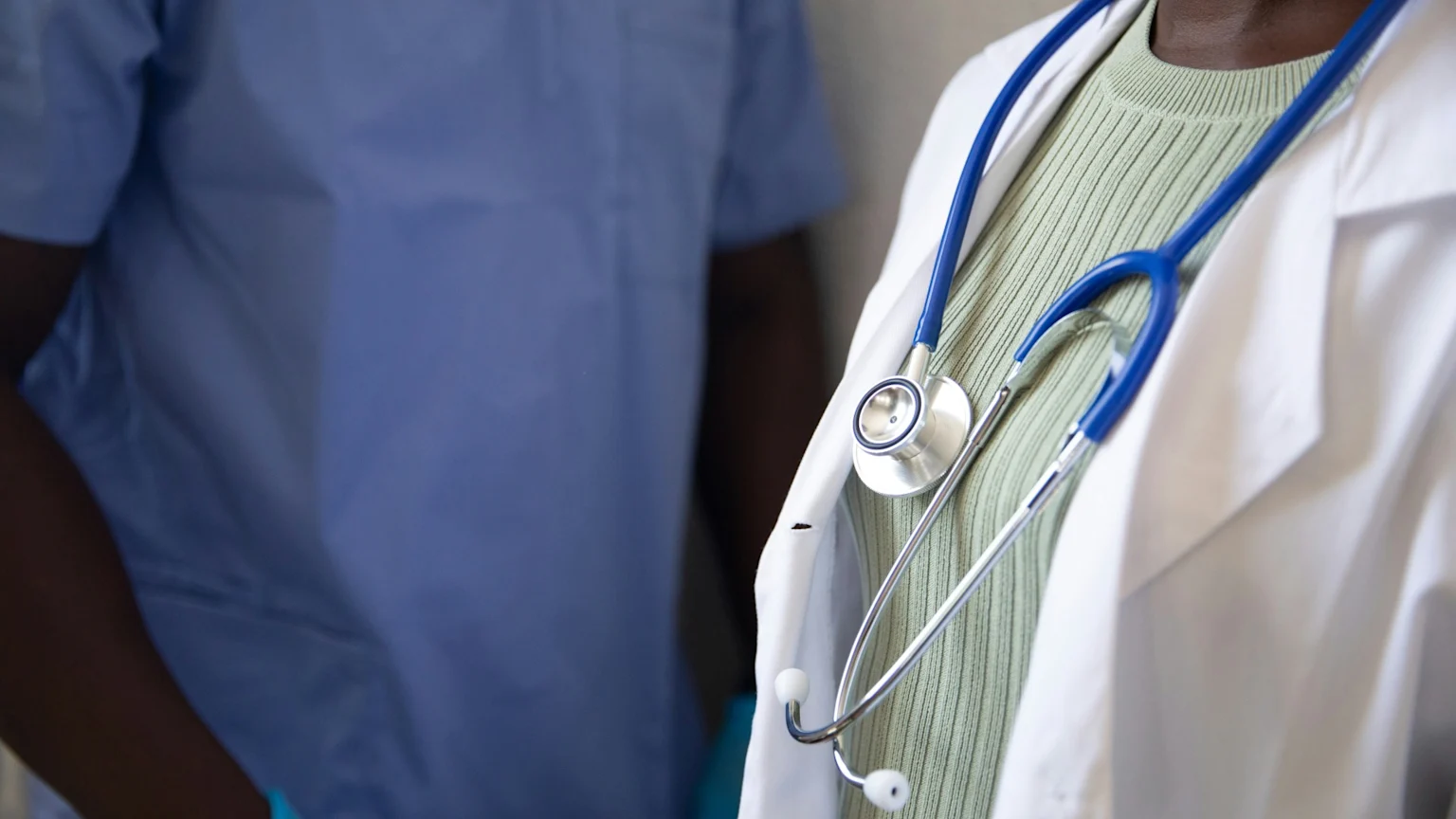 Close-up of two medical professionals, one wearing a white lab coat and a stethoscope, the other in blue scrubs.