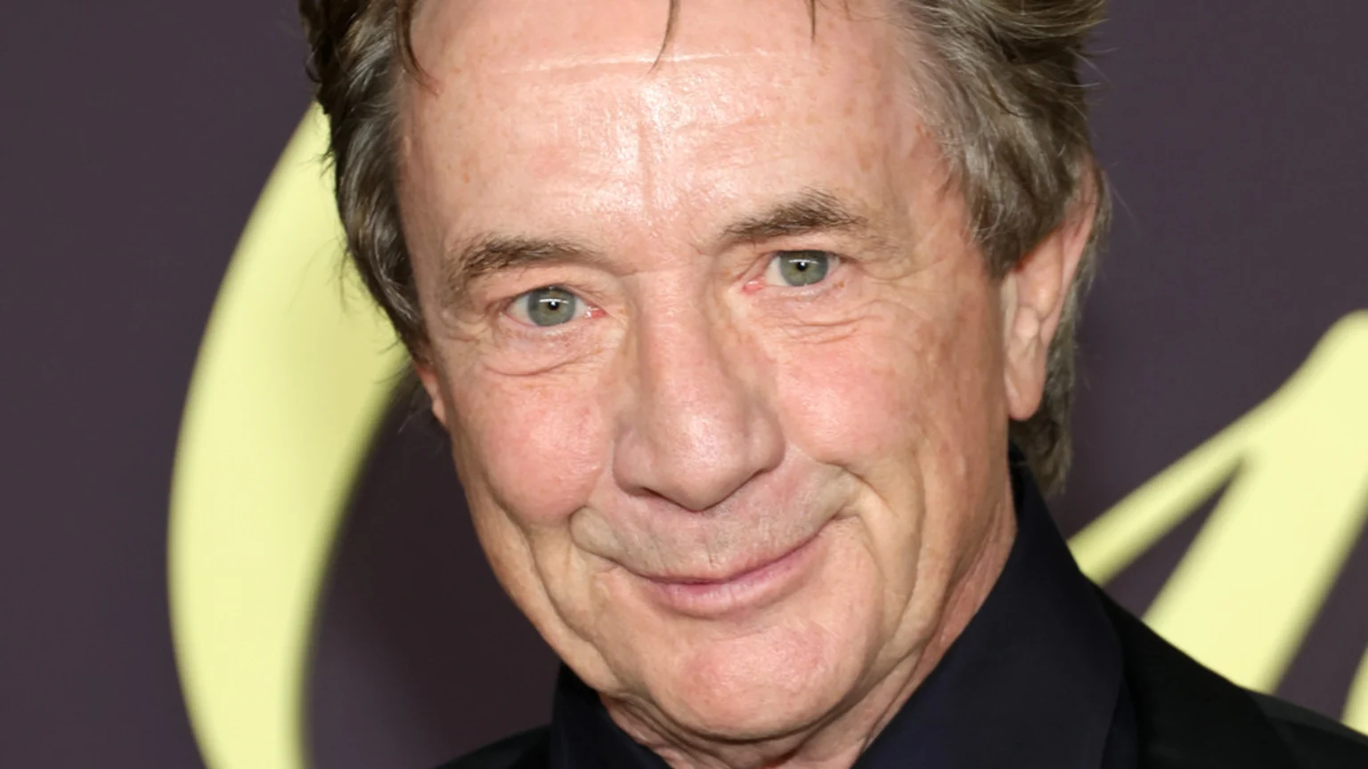 A close-up shows Martin Short smiling gently at the camera, with light eyes and grey-brown hair, wearing a dark collared shirt against a blurred dark and yellow background.