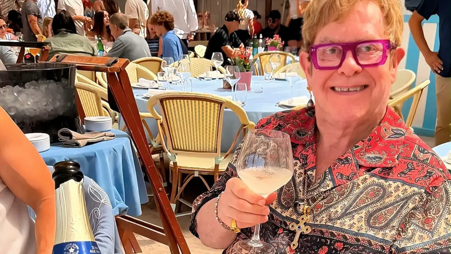 Elton John smiles, holding a glass of sparkling drink, wearing purple glasses, a colorful patterned shirt, and a cross pendant, at an outdoor cafe.