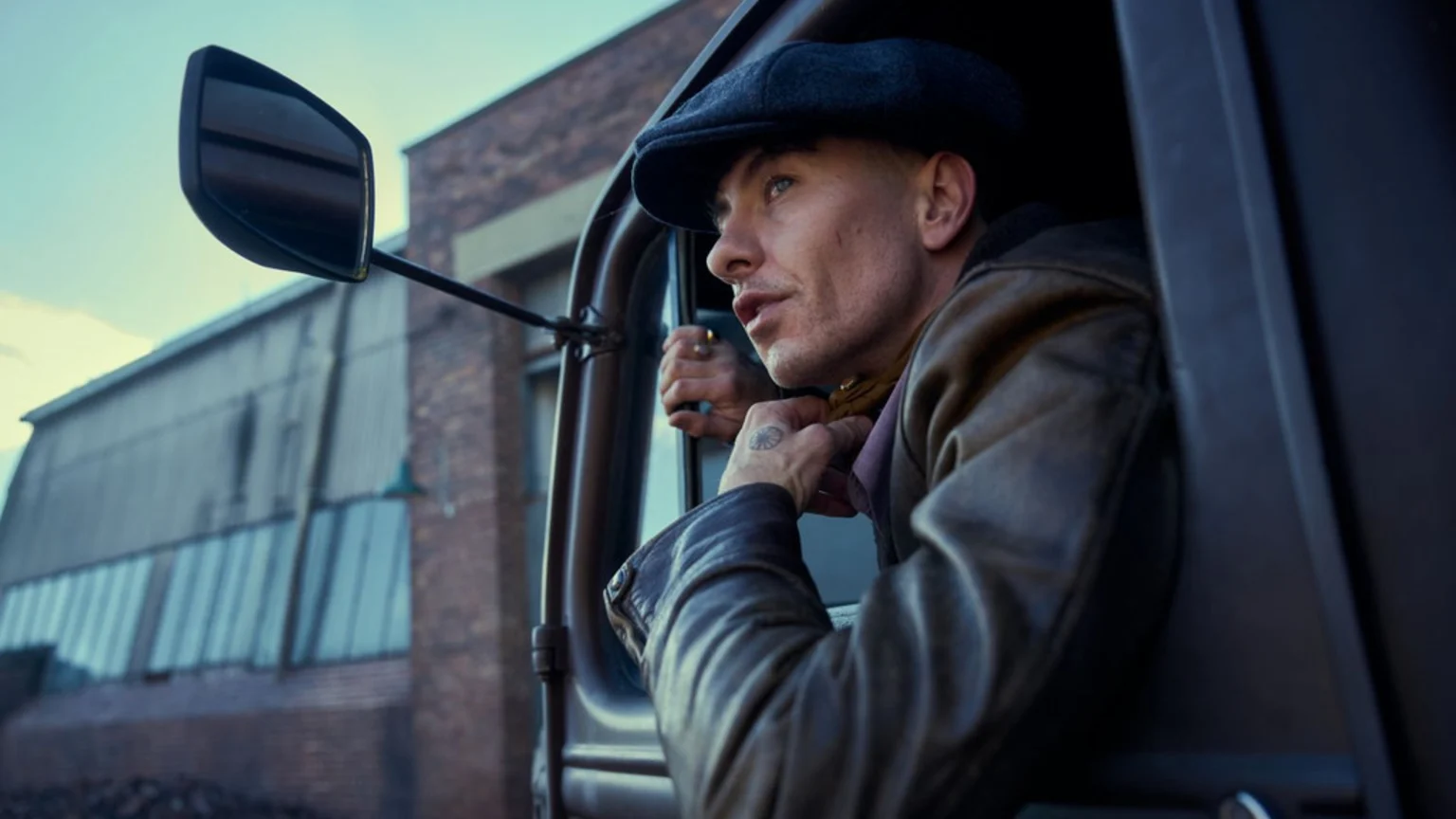 A man in a dark cap and brown jacket leans out of a vehicle window, looking left, with an industrial building in the blurred background.